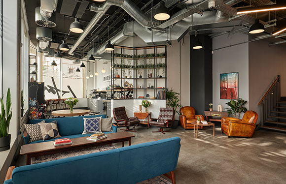 A modern lounge with blue velvet sofas, leather armchairs, and industrial exposed ceilings.