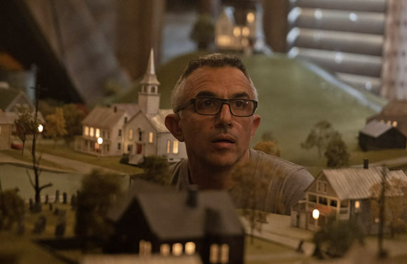 A man with glasses looks intently at a detailed miniature model of a small town with houses and a church.