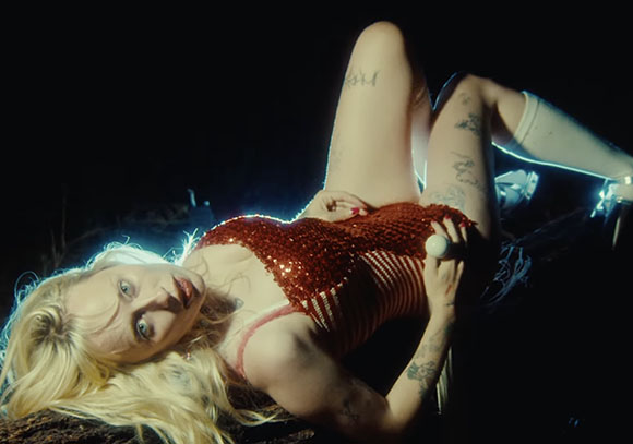 A blonde woman in a red sequined bodysuit lies on her back against a dark background.