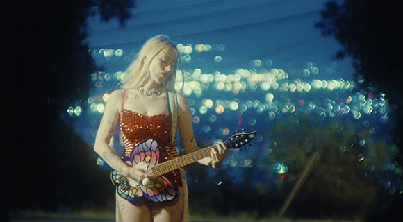 A blonde woman in a red sequined bodysuit plays an electric guitar at night with city lights in the background.