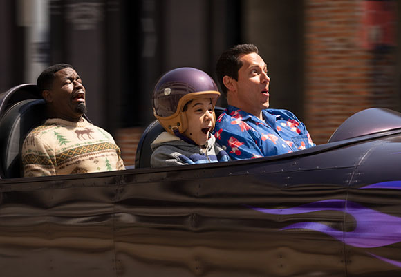 A man, a child in a purple helmet, and another man riding a roller coaster with excited expressions.