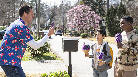 A man holds up a phone to photograph two boys holding large ice cream cones outdoors.