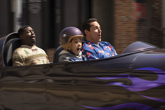 A man, a child in a helmet, and another man riding a dark amusement park vehicle.