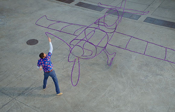 A person in a blue patterned shirt draws a large purple outline of an airplane on a concrete ground.