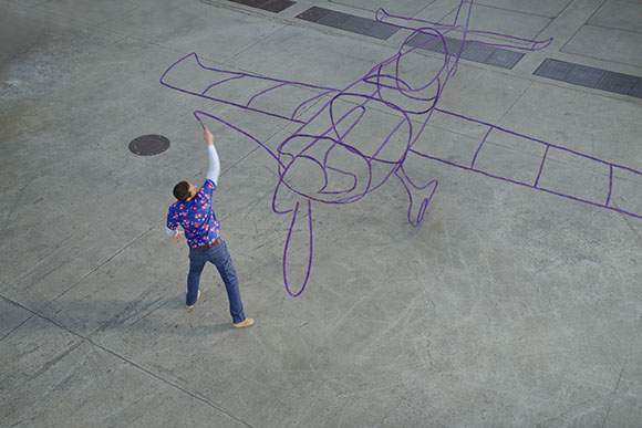 A person in a blue patterned shirt draws a large purple outline of an airplane on a concrete ground.