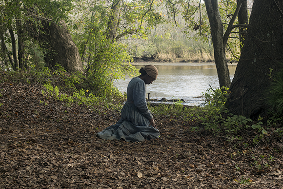 A woman in a long blue dress kneels on a bed of fallen leaves near a riverbank in a forest.