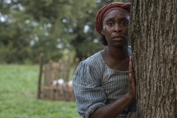 A woman in period clothing peeks cautiously from behind a large tree trunk in a grassy field.