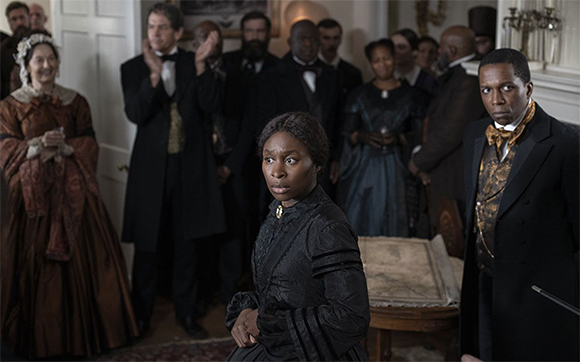 A Black woman in a black dress looks sideways while a group of people in period clothing stand behind her.