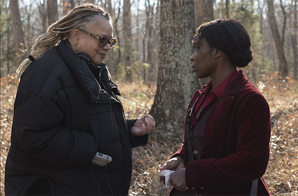 A woman with blonde braids in a black puffer coat talks to a woman in a red velvet jacket in a forest.
