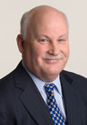 A smiling man with a bald head, wearing a dark suit, light blue shirt, and blue patterned tie.