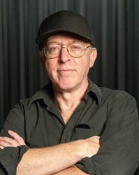 A man with glasses and a black baseball cap poses with arms crossed against a dark curtain.