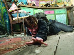A young man with glasses crawls on a wooden floor, reaching his hand toward the camera.