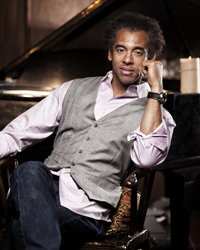 A man with curly hair wearing a grey vest and pink shirt sits in a chair next to a piano.