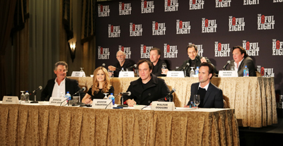 Cast members sit behind a long table with microphones in front of a black backdrop with white text.