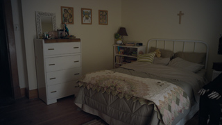A dimly lit bedroom featuring a white dresser, a bed with a patterned quilt, and a cross on the wall.