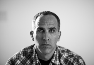 Black and white close-up portrait of a man looking directly at the camera with a neutral expression.