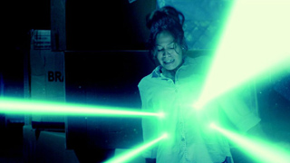 A woman in a white shirt stands in a dark room as bright green light beams cross in front of her.