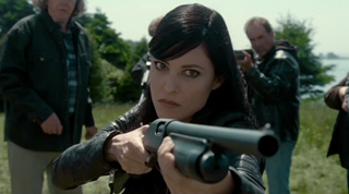 A woman with dark hair aims a shotgun forward in a grassy field with men behind her.