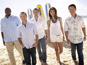 Five people in casual summer clothing standing on a sandy beach with surfboards in the background.