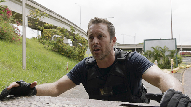 A man wearing a black tactical vest looks intensely to the side against a background of a highway overpass.