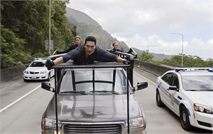A man hangs onto the roof rack of a dark SUV while being pursued by two white police cars on a highway.