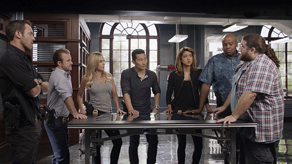 Seven diverse people stand in a circle around a large metal table in a modern office.