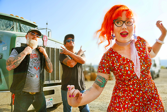 A woman with bright orange hair and a red dress poses in front of two men standing by a large green truck.