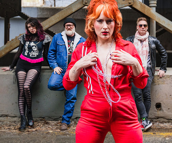 A woman in a red jumpsuit stands in front of three people leaning against a concrete wall.