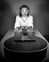 Black and white portrait of a man with long hair and white glasses holding an acoustic guitar.