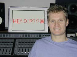 A man in a purple shirt smiles in front of a computer monitor displaying the word HEADROOM in a studio setting.
