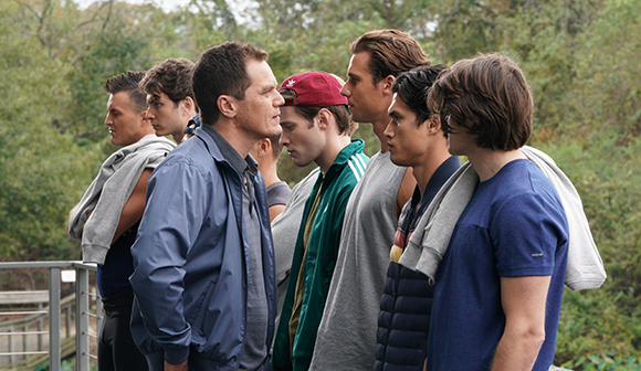 A line of seven young men standing outdoors against a backdrop of green trees.