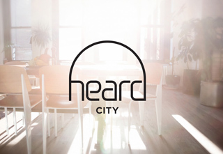Black logo with the text 'heard CITY' centered over a bright, sunlit interior with chairs and plants.