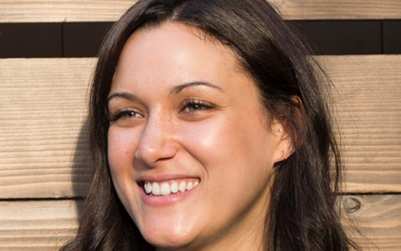 A smiling woman with dark hair looking slightly to the side against a wooden background.