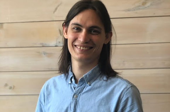 A smiling man with shoulder-length brown hair wearing a light blue button-down shirt against a wooden wall.