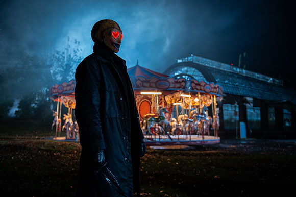 A person wearing a dark coat and a mask with glowing red heart eyes stands before a lit carousel at night.