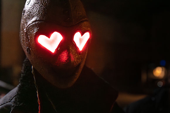 A person in a dark, textured mask with two glowing red heart-shaped eyes.