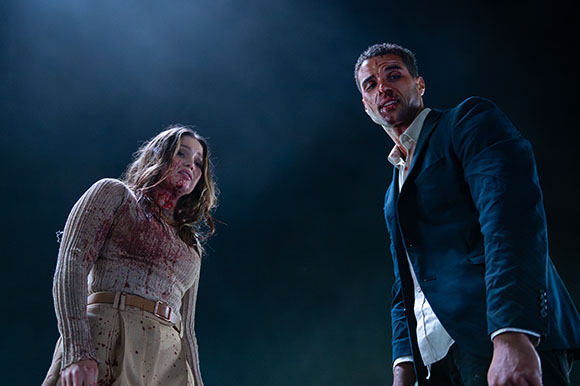 A blood-stained woman in a beige sweater and a man in a dark suit stand against a dark, smoky backdrop.
