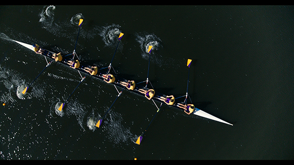 Aerial view of eight rowers in a long white rowing shell on dark water, creating white splashes with their oars.
