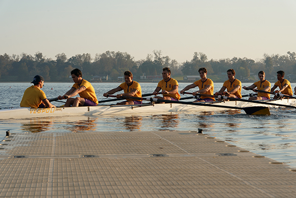 A rowing crew of eight athletes in yellow shirts rowing a long white boat on calm water at sunrise.