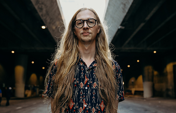 A man with long blonde hair and black-rimmed glasses wearing a patterned shirt under a concrete bridge.