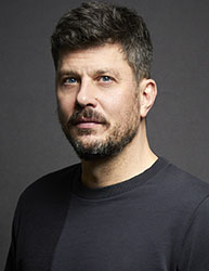 A man with short dark hair, blue eyes, and a salt-and-pepper beard looking toward the camera against a dark background.