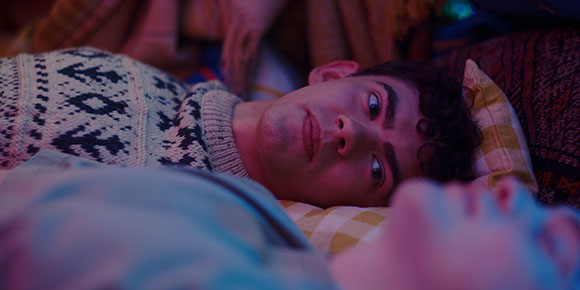 A young man with dark curly hair lies on a pillow, looking upward with a serious expression.