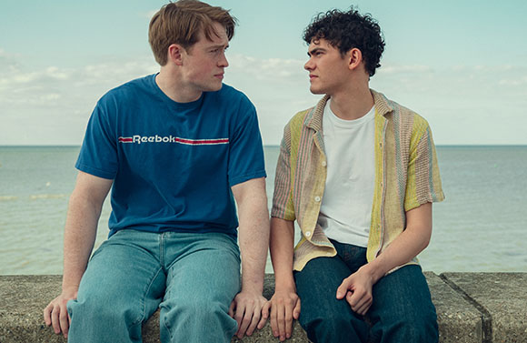 Two young men sit on a stone wall by the ocean, looking at one another.
