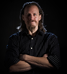 A man with long hair and a goatee wearing a dark striped shirt, posing with arms crossed against a black background.