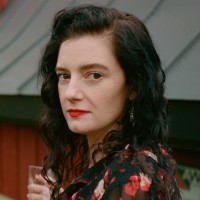 A woman with dark wavy hair and red lipstick looking toward the camera against a blurred background.