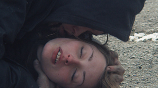 A person in a dark hoodie leans over a person lying on a gravel surface with their eyes closed.