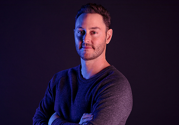 A man with short dark hair and a beard wearing a dark sweater, looking at the camera against a dark background.
