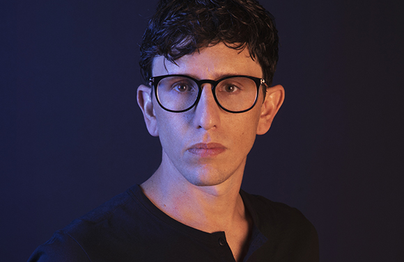 A man with dark curly hair and black-rimmed glasses looks directly at the camera against a dark blue background.