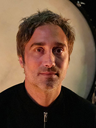 A man with short, messy brown hair and light stubble looks directly at the camera.
