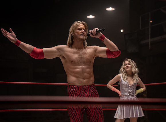 A shirtless man with long blonde hair holds a microphone in a wrestling ring next to a woman in a silver dress.